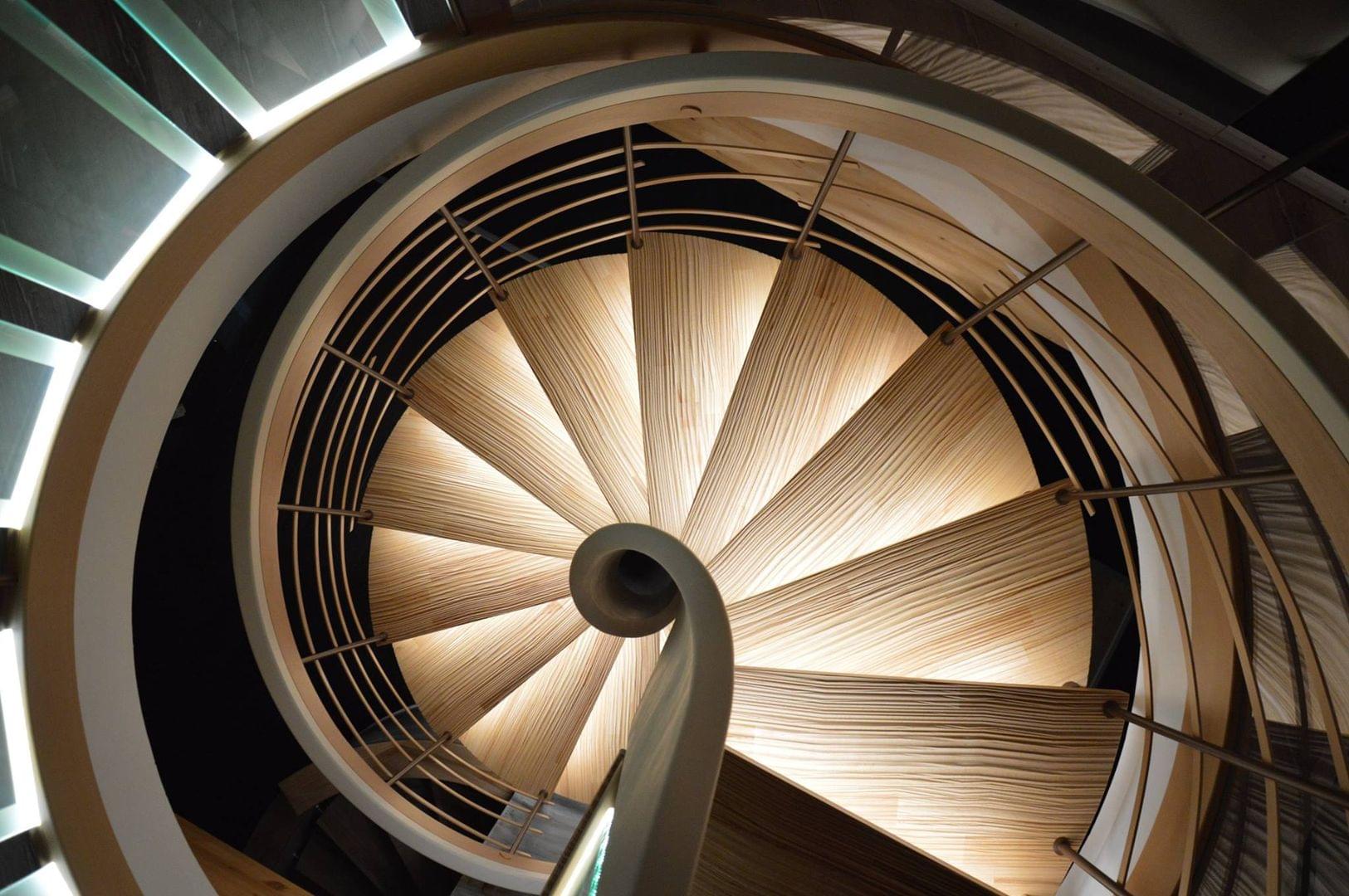 Escalier courbe de forme colimaçon ou hélicoîdale. Main-courante débillardé en lamellé collé et avec une finition bi-teinte. Marches avec usinage de surface senzu. Rampe avec balustres acier laqué et "feuillage" bois..Eclairage sous marches