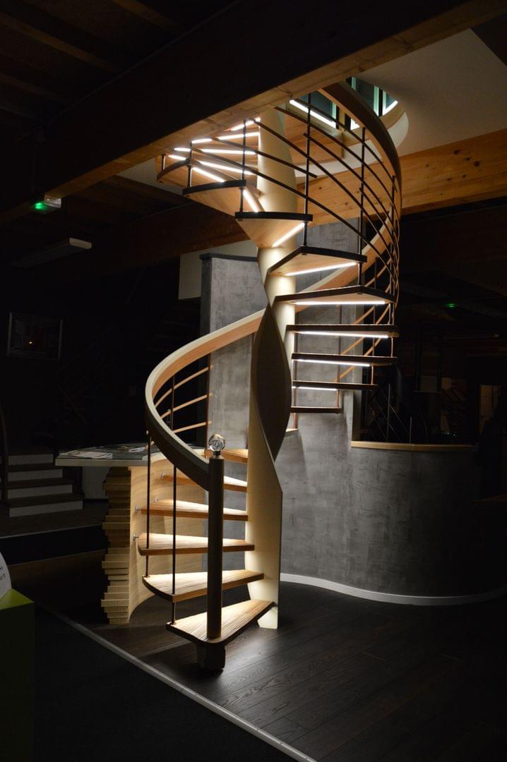 Escalier courbe de forme colimaçon ou hélicoîdale. Main-courante débillardé en lamellé collé et avec une finition bi-teinte. Marches avec usinage de surface senzu. Rampe avec balustres acier laqué et "feuillage" bois..Eclairage sous marches